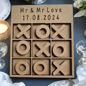 May include: A wooden tic-tac-toe game with the text "Mr & Mr Love 17.08.2024" on the top. The game is made of light brown wood and has a natural finish.