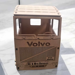 May include: A wooden Volvo truck model with etched details. The truck features the Volvo logo, F12 Volvo, and the names "Mr & Mrs Benson" with the date "21.08.2020". The model has a rectangular shape with a detailed front and a roof.