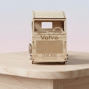 May include: A light brown wooden model of a Volvo lorry. The model features etched details, including the Volvo logo and the license plate "TIF XO". The lorry sits on a light-coloured wooden surface.