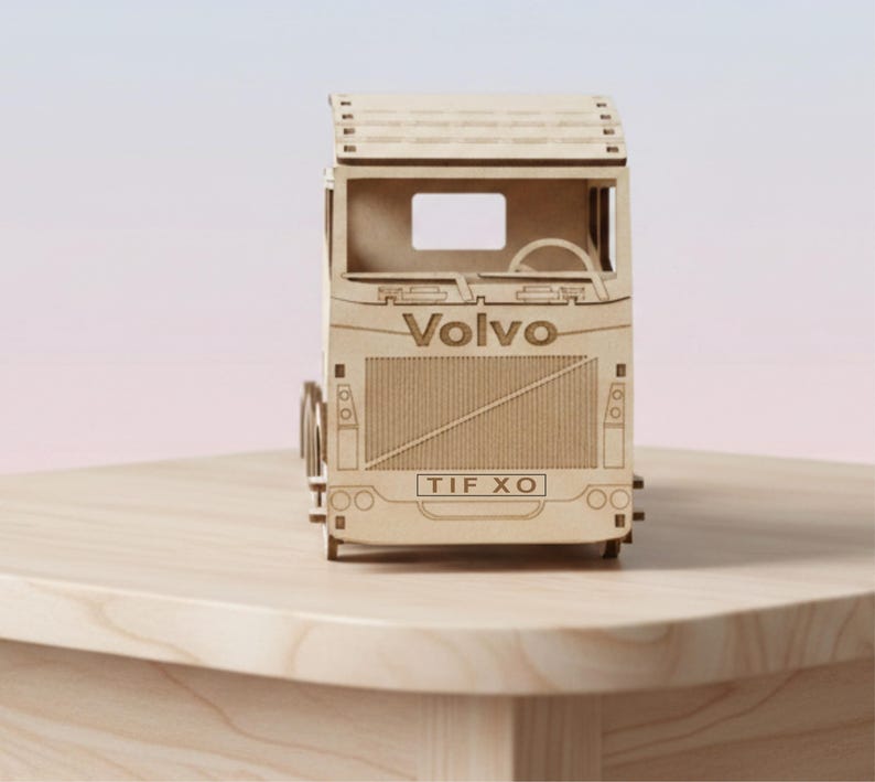 May include: A light brown wooden model of a Volvo lorry. The model features etched details, including the Volvo logo and the license plate "TIF XO". The lorry sits on a light-coloured wooden surface.
