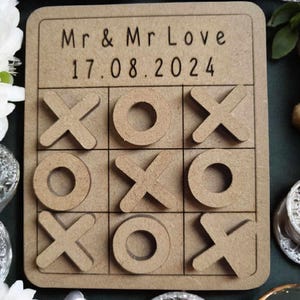 May include: A wooden tic-tac-toe game with the text "Mr & Mr Love 17.08.2024" on the top. The game board is brown and has a white background.