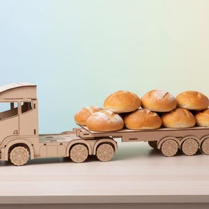 May include: A wooden toy truck transporting a stack of fresh, golden-brown bread rolls. The truck is made of light-coloured wood and has detailed cutouts. The rolls are round and appear freshly baked, suggesting a bakery or food theme.