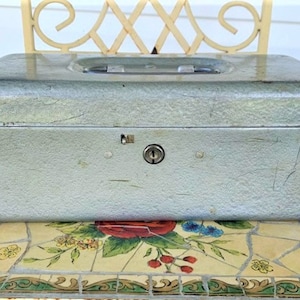 May include: A vintage metal cash box with a silver finish and a keyhole. The box has a textured surface and a handle on the top.