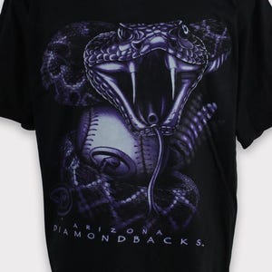 May include: Black t-shirt featuring a purple graphic of a snake with its mouth open, fangs bared, over a baseball. The text "ARIZONA DIAMONDBACKS" is printed in white below the graphic. The shirt is displayed on a mannequin.