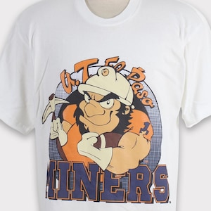May include: White t-shirt with a cartoon mascot wearing a hard hat and holding a pickaxe. The mascot is surrounded by a circle with the text "U.T. El Paso Miners".