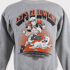 Puede incluir: Sudadera gris con un jugador de fútbol de dibujos animados y un perro corriendo. El texto "LET'S GO BOWLING" está impreso en naranja en la parte posterior de la sudadera.