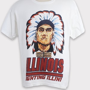 May include: White t-shirt with a Native American chief illustration wearing a feathered headdress. The word "ILLINOIS" is printed in red block letters, with "FIGHTING ILLINI" below. The shirt is displayed on a mannequin.