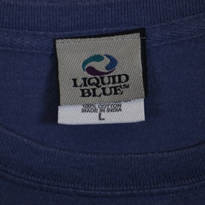 Puede incluir: Una etiqueta de camiseta azul de la marca Liquid Blue con texto blanco que dice "100% Algod&oacute;n Hecho en India" y una etiqueta de talla "L".