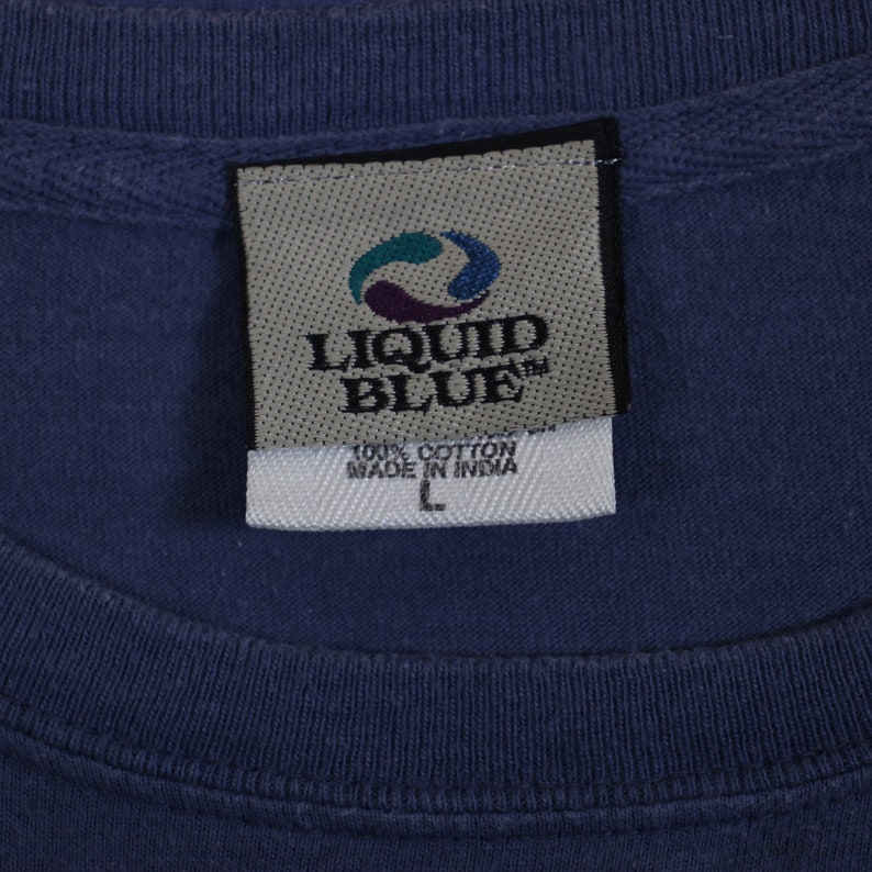 Puede incluir: Una etiqueta de camiseta azul de la marca Liquid Blue con texto blanco que dice "100% Algod&oacute;n Hecho en India" y una etiqueta de talla "L".