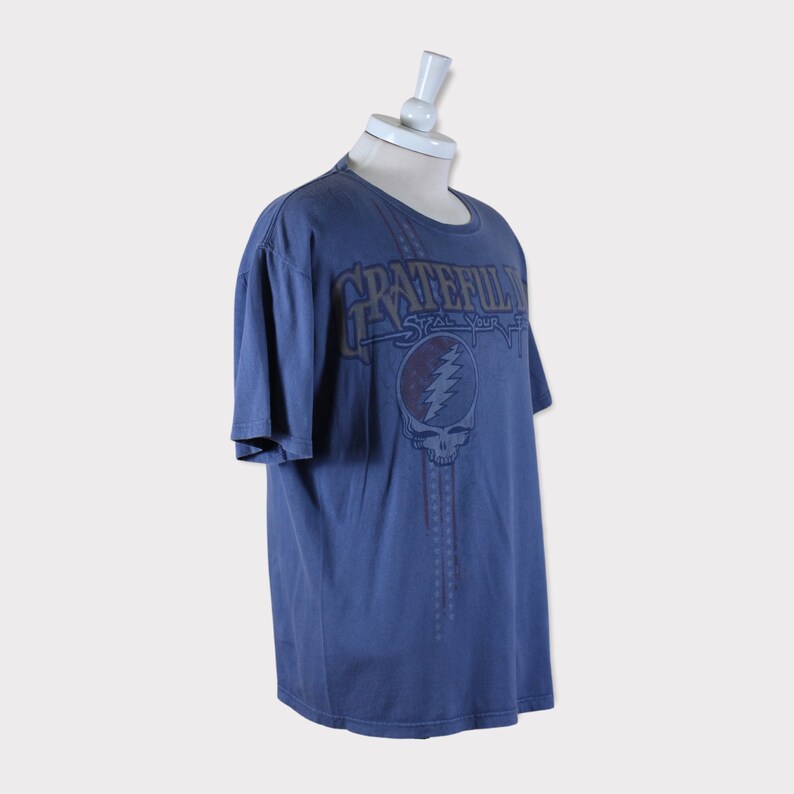 Puede incluir: Una camiseta azul con un estampado descolorido del logotipo de Grateful Dead Steal Your Face. El logotipo es de color marr&oacute;n, rojo y blanco.