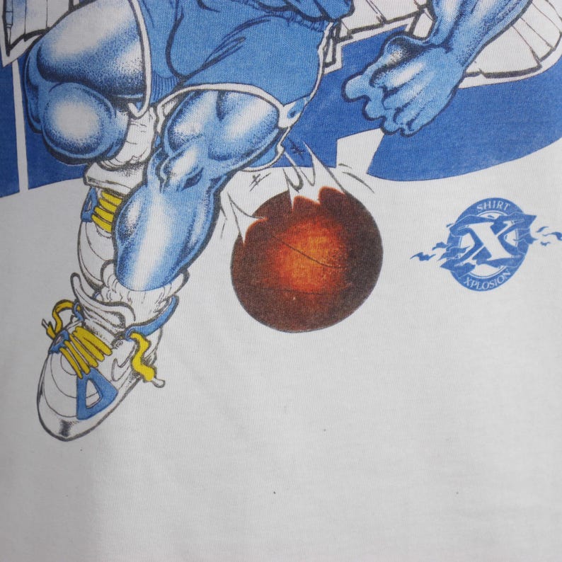 May include: White t-shirt featuring a cartoon illustration of a basketball player in blue and white. The player is dribbling a basketball with a fiery effect. The shirt has the text "Shirt Xplosion".
