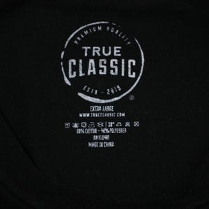 May include: A black tag with white text that reads "Premium Quality True Classic Estb - 2019" with a circle around the text. The tag also includes information about the size, fabric content, and origin of the garment.