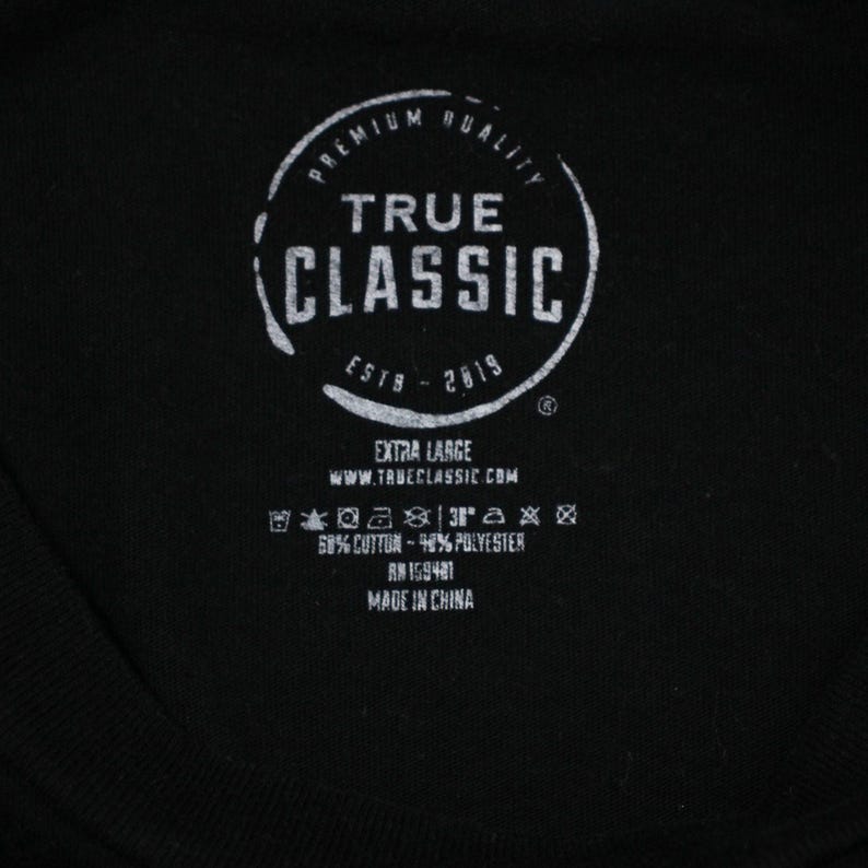 May include: A black tag with white text that reads "Premium Quality True Classic Estb - 2019" with a circle around the text. The tag also includes information about the size, fabric content, and origin of the garment.