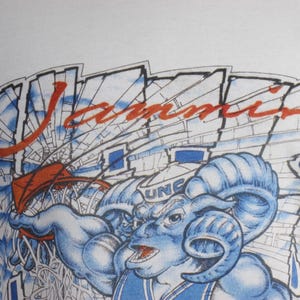 May include: White t-shirt featuring a blue ram mascot with the text "UNC" and the word "Jammin" in red. The design includes a basketball net and a stadium background.