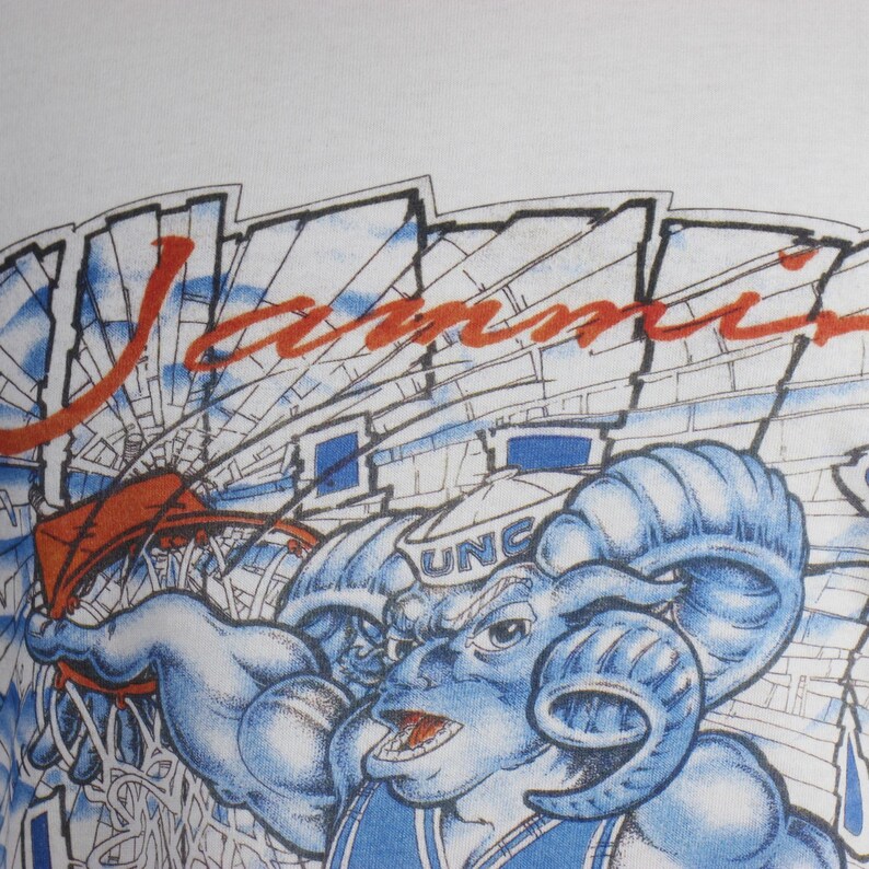 May include: White t-shirt featuring a blue ram mascot with the text "UNC" and the word "Jammin" in red. The design includes a basketball net and a stadium background.