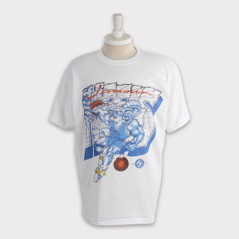 May include: White t-shirt featuring a blue and red graphic of a ram playing basketball. The word "Summit" is printed above the ram. The ram is wearing a blue basketball uniform and holding a basketball.