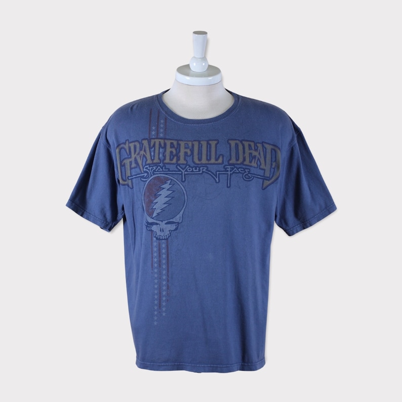 Puede incluir: Una camiseta azul con un estampado descolorido del logotipo de Grateful Dead y el texto "Steal Your Face".
