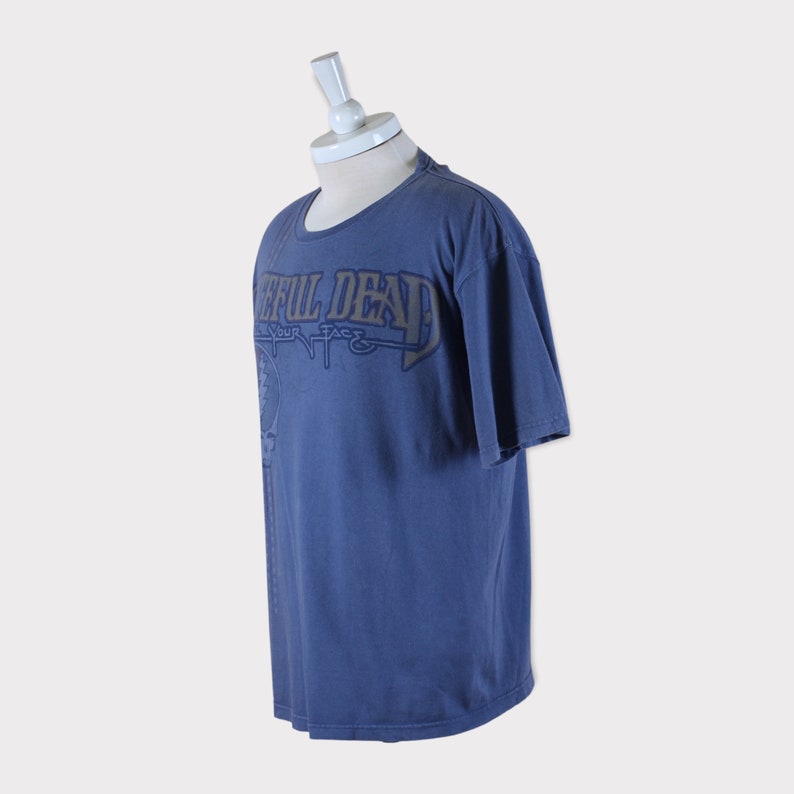 Puede incluir: Una camiseta azul con un estampado gr&aacute;fico descolorido de "Grateful Dead" en una fuente estilizada. El texto "Your Face" est&aacute; impreso debajo del nombre de la banda.