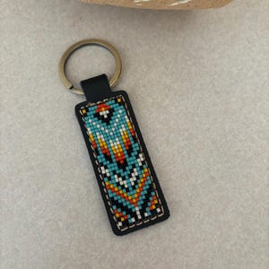 May include: A black leather keychain with a colorful beaded design. The design features geometric shapes in shades of blue, orange, white, and black.