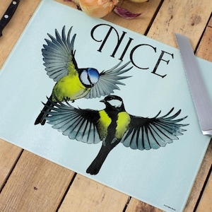 May include: A pale blue glass chopping board decorated with two birds in flight. The word "NICE" is printed in black script above the birds. A knife is visible on the right side of the board.