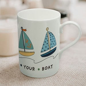 Puede incluir: Taza de cerámica blanca con un diseño de velero azul y amarillo. La taza tiene el texto "Your Boat" en la parte inferior.