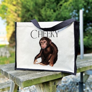 May include: A white tote bag with black trim and handles. It displays a brown chimpanzee illustration and the word "CHEEKY" in black. The bag is resting on a wooden bench.