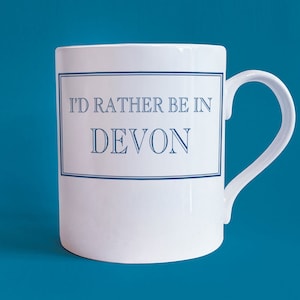 Könnte beinhalten: Weiße Keramiktasse mit einem blauen Rand um den Text "I'd rather be in Devon".