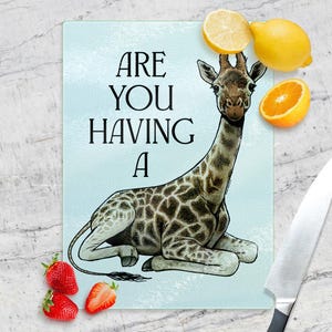 Könnte beinhalten: Ein rechteckiges Glas-Schneidebrett mit einer Giraffen-Illustration und dem Text "ARE YOU HAVING A". Die Giraffe ist braun und weiß mit einem langen Hals. Zitronen, eine Orangenscheibe und Erdbeeren liegen neben dem Brett. Rechts ist ein Messer.