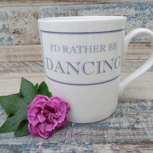 May include: White ceramic mug with a blue border and the text "I'd rather be dancing" in blue lettering. A pink carnation and green leaves are in the foreground.
