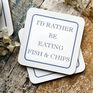Può includere: Due sottobicchieri quadrati bianchi con un bordo blu. Il sottobicchiere superiore ha il testo "I'd rather be eating fish & chips" in lettere blu.