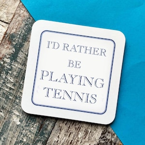 Peut inclure: Sous-verre blanc avec une bordure bleue et le texte "I'd rather be playing tennis" en bleu.