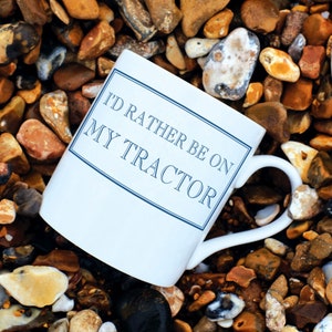 Könnte beinhalten: Weiße Keramiktasse mit blauem Text, der "Ich wäre lieber auf meinem Traktor" lautet. Die Tasse steht auf einem Bett aus kleinen, bunten Steinen.