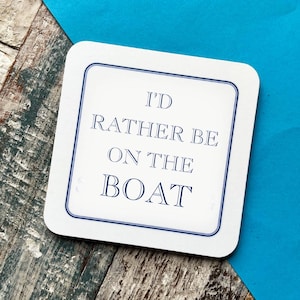 May include: White square coaster with a blue border and the text "I'd rather be on the boat" in blue.