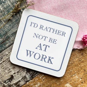 May include: Square white coaster with a navy blue border and the text "I'D RATHER NOT BE AT WORK" in navy blue. The coaster is on a rustic wooden surface with a pink cloth and dried flowers in the background.