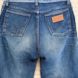 May include: Close-up of the rear of blue denim jeans. The jeans feature two back pockets with a distinctive "W" stitch. A brown leather patch with the "Wrangler" logo is visible on the right pocket.