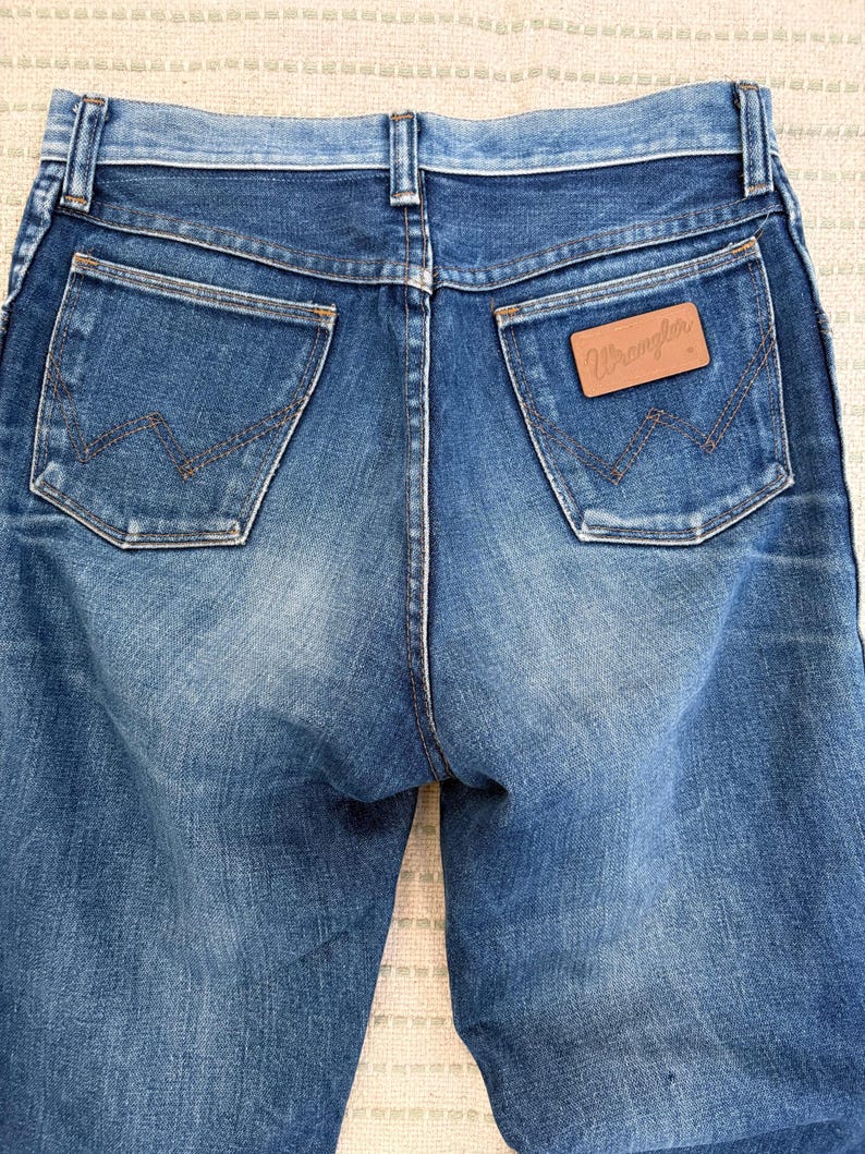 May include: Close-up of the rear of blue denim jeans. The jeans feature two back pockets with a distinctive "W" stitch. A brown leather patch with the "Wrangler" logo is visible on the right pocket.