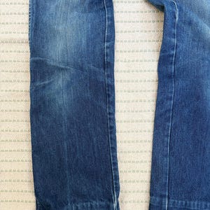 May include: Close-up of denim jeans in a medium blue wash. The jeans have a straight leg cut and a classic five-pocket design. The fabric shows subtle fading and wear, adding to the vintage aesthetic. The image highlights the texture and colour variations of the denim.