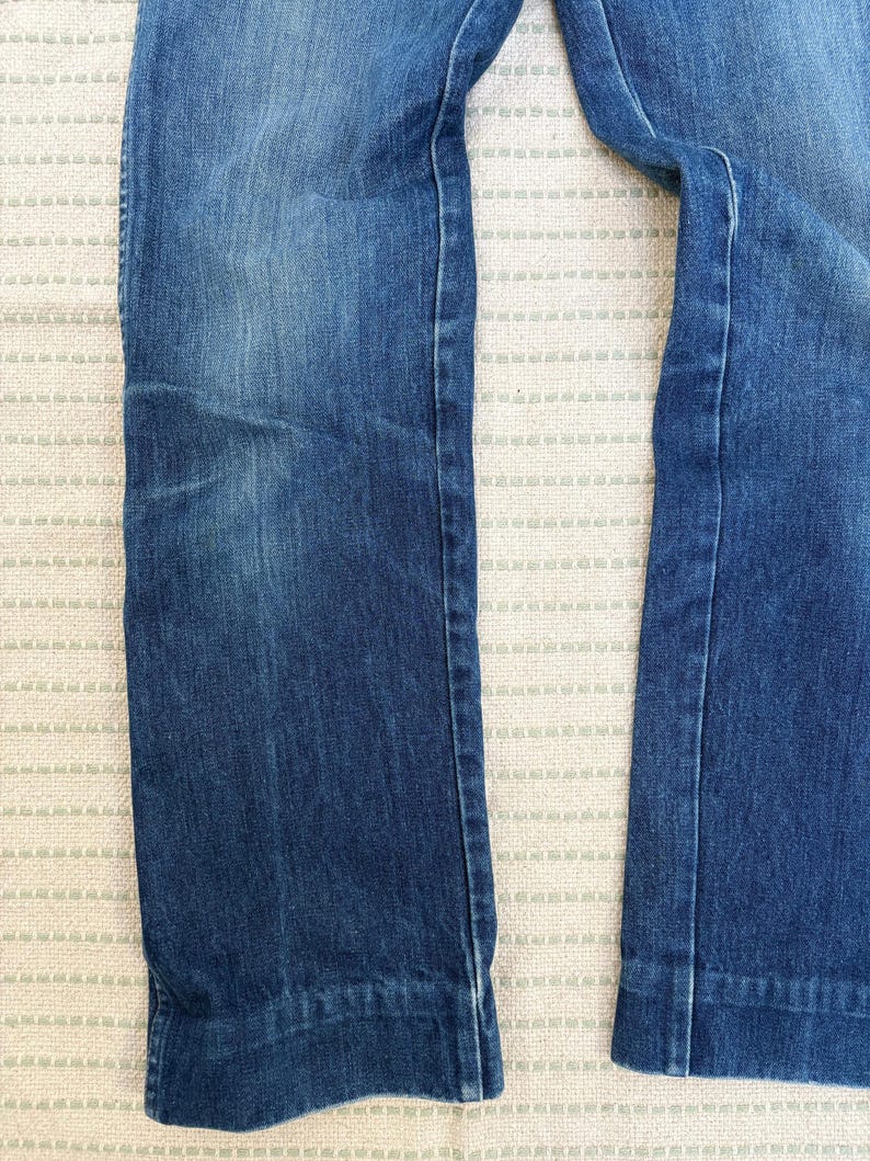 May include: Close-up of denim jeans in a medium blue wash. The jeans have a straight leg cut and a classic five-pocket design. The fabric shows subtle fading and wear, adding to the vintage aesthetic. The image highlights the texture and colour variations of the denim.