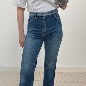 May include: Blue denim jeans with a straight-leg cut. The jeans feature a high waist and button closure. The denim has a faded wash, giving a vintage appearance. The jeans are worn with a white t-shirt.