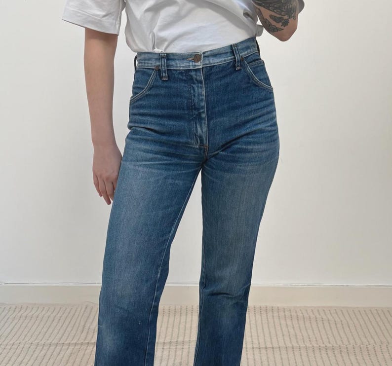 May include: Blue denim jeans with a straight-leg cut. The jeans feature a high waist and button closure. The denim has a faded wash, giving a vintage appearance. The jeans are worn with a white t-shirt.