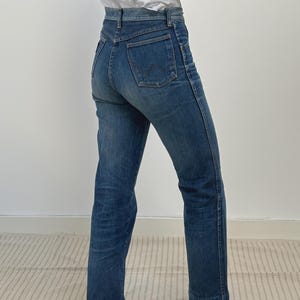 May include: Blue denim jeans with a high waist and a straight leg. The jeans have a classic five-pocket design and a button and zip closure. The jeans are worn with a white t-shirt and dark shoes.