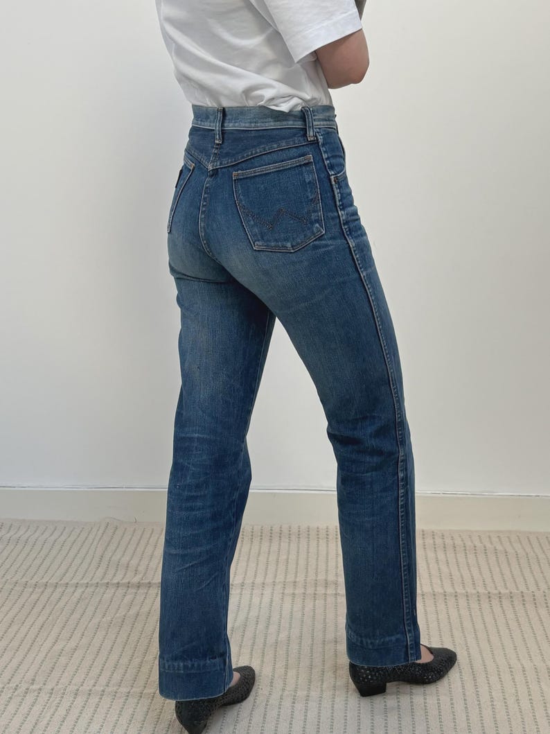 May include: Blue denim jeans with a high waist and a straight leg. The jeans have a classic five-pocket design and a button and zip closure. The jeans are worn with a white t-shirt and dark shoes.