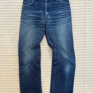 May include: Vintage blue denim jeans with a high waist and straight leg. The jeans feature a classic five-pocket design and a button and zip closure. The denim has a faded wash with some wear and tear.