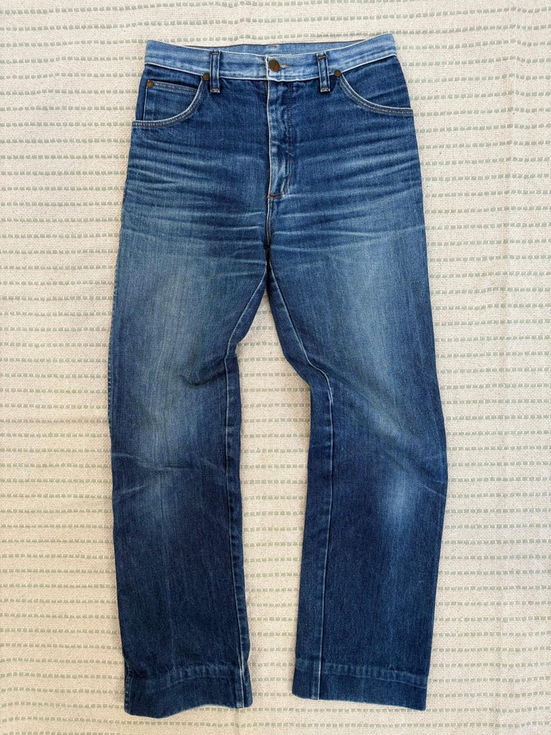 May include: Vintage blue denim jeans with a high waist and straight leg. The jeans feature a classic five-pocket design and a button and zip closure. The denim has a faded wash with some wear and tear.