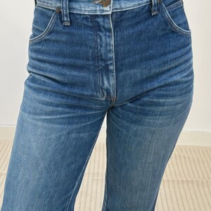 May include: Close-up of blue denim jeans. The jeans feature a button closure, belt loops, and front pockets. The denim has a faded look, with lighter areas on the thighs and knees. The jeans are a classic, straight-leg style.
