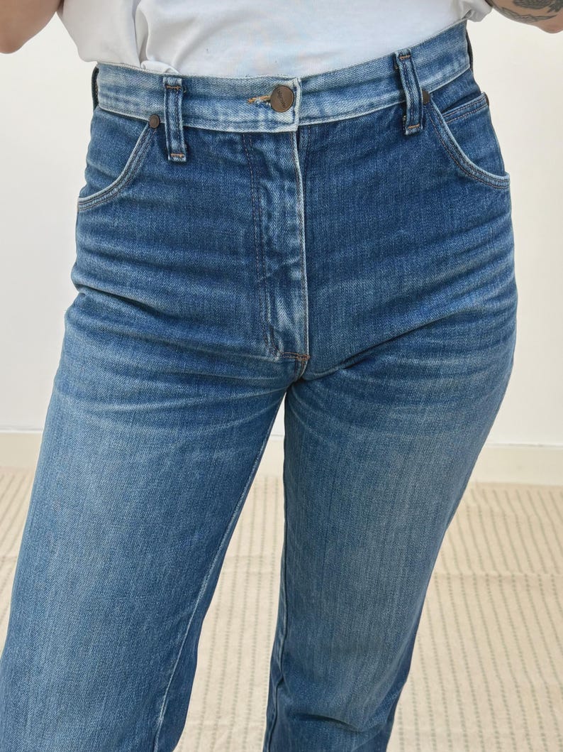 May include: Close-up of blue denim jeans. The jeans feature a button closure, belt loops, and front pockets. The denim has a faded look, with lighter areas on the thighs and knees. The jeans are a classic, straight-leg style.