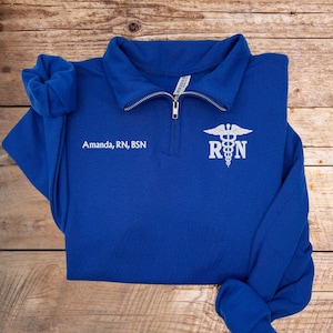 May include: Royal blue quarter-zip sweatshirt with white embroidered text and medical symbol. The text reads "Amanda, RN, BSN" on the left chest and "RN" with a caduceus symbol on the right chest. The sweatshirt is folded on a wooden surface.