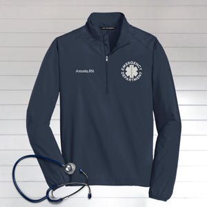 May include: Navy blue quarter-zip jacket with the text "Antonio, RN" and "Emergency Department" with a medical symbol. A stethoscope is in the foreground. The jacket has a zipper and is made of a lightweight material.
