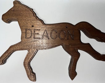 May include: A dark brown wooden wall sign shaped like a galloping horse with the name DEACON engraved in capital letters across the center. The cutout silhouette features a smooth finish and two small circular holes for mounting purposes.