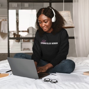 K&ouml;nnte beinhalten: Eine Frau tr&auml;gt ein schwarzes Sweatshirt mit dem Text "Soft Strong Black Woman" in rosa Schrift, sitzt auf einem Bett und benutzt einen Laptop. Sie tr&auml;gt Kopfh&ouml;rer und hat lockiges schwarzes Haar.